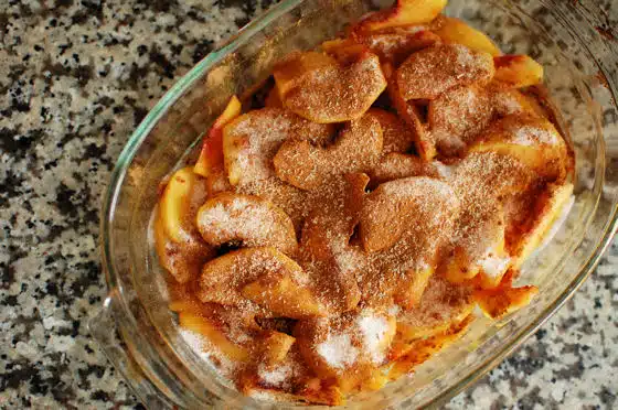 A glass baking dish filled with sliced apples covered in a mixture of cinnamon and sugar, resting on a speckled countertop.