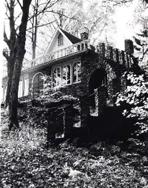 Black-and-white image of a large stone house with arched windows and a turret, surrounded by trees and foliage.