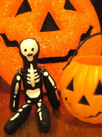 A small black and white skeleton figurine sits in front of a glittery orange jack-o'-lantern backdrop, next to a pumpkin-shaped container.