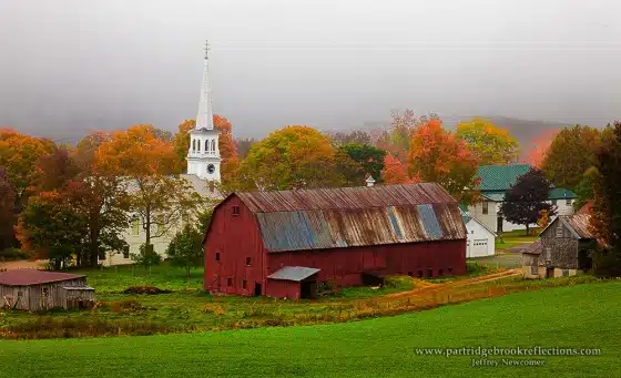A red barn sits in a green field in front of a white church with a tall steeple surrounded by autumn foliage under a foggy sky.