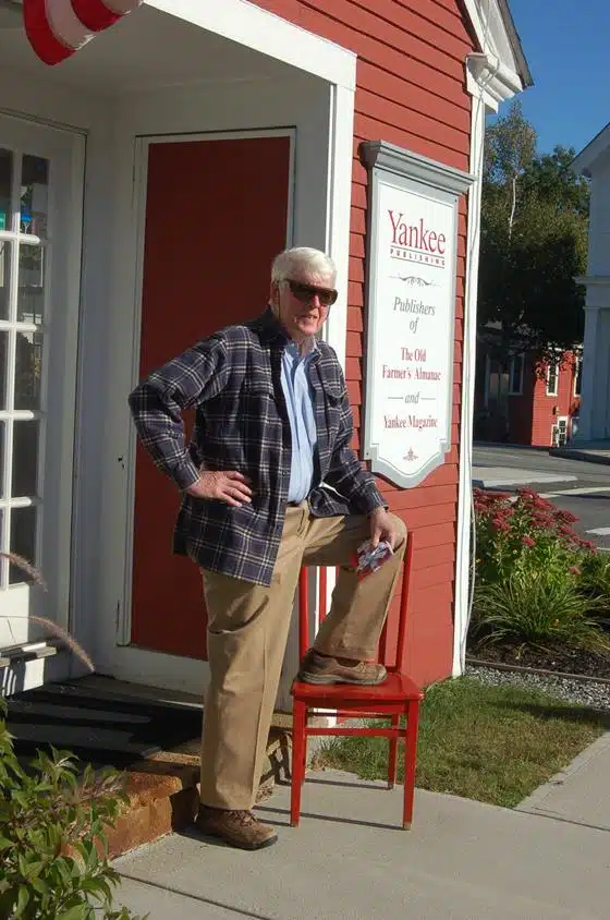 A man with white hair and sunglasses stands with one foot on a red chair in front of a red and white building that has a sign for Yankee Publishing.
