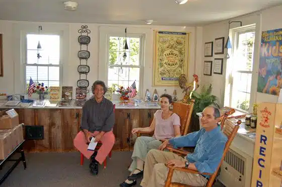 Three people are seated indoors, two on rocking chairs. The room has windows, bright posters on the walls, and a countertop with various items, including small American flags and decorative objects.