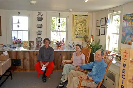 Three people sitting in a brightly lit room with various posters, boxes, certificates, and a bookshelf. One person is seated on the left in red pants, and the other two are on rocking chairs.
