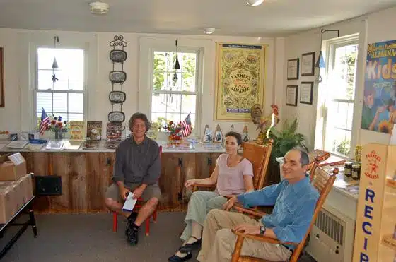 Three people sitting inside a room with various memorabilia, posters, and decorations on the walls. Two of them are seated on wooden rocking chairs while one is seated on a stool.