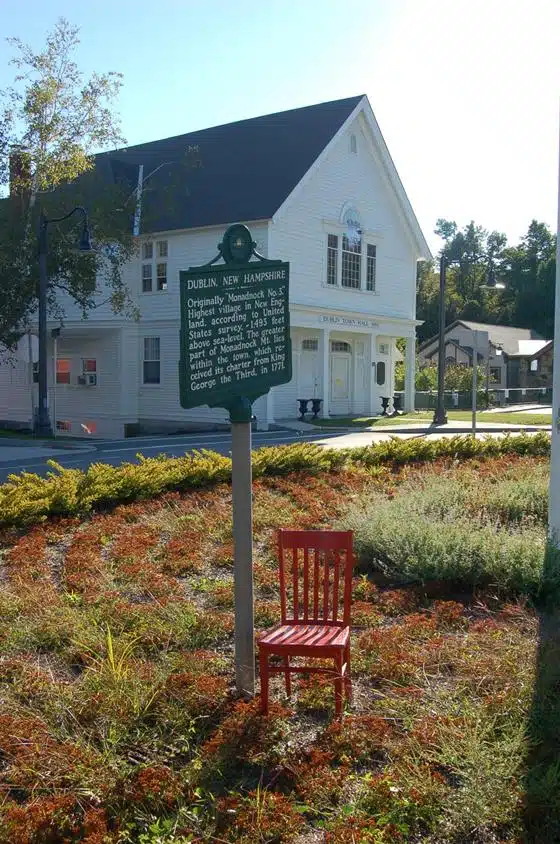 A historical marker in front of a building in Dublin, New Hampshire, with a red chair placed next to the marker. The area is landscaped with sparse ground cover and bushes.