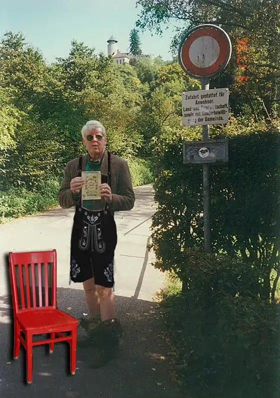 A man in traditional attire holds a booklet while standing on a path next to a sign and a red chair in a scenic outdoor setting.