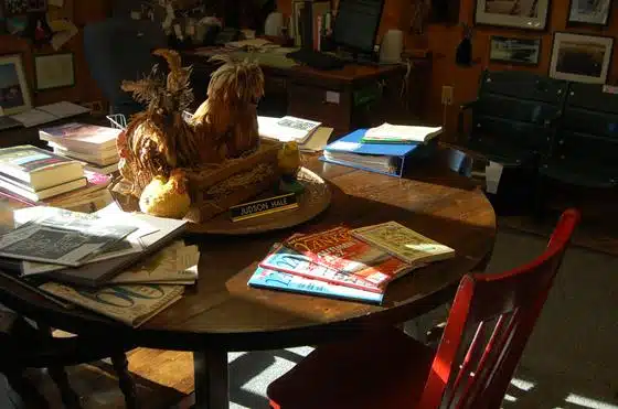 A cluttered round table with books, papers, and a decorative rooster centerpiece. A red chair is pushed against the table. Office supplies and framed pictures are visible in the background.