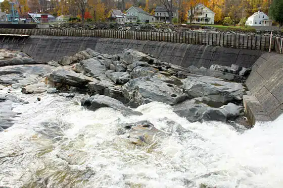 A rocky dam with water flowing over the rocks and cascading down. Houses and trees with fall foliage are visible in the background.