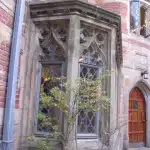 A gothic-style window with intricate stonework and leaded glass panes on a brick building, accompanied by a small tree in the foreground.