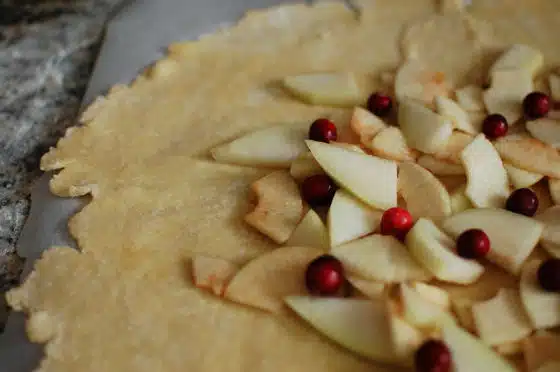 Unbaked pie crust topped with sliced apples and cranberries, laid out on a floured surface.