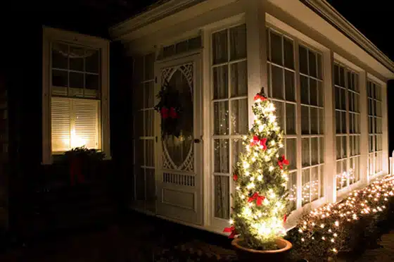 A small tree decorated with lights and red ribbons stands next to a front door adorned with a wreath. The house's enclosed porch is lit up, and string lights line the base of the wall.