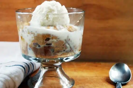 A glass dessert dish containing tiramisu topped with a scoop of vanilla ice cream, placed next to a napkin and a spoon on a wooden surface.