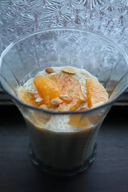 A glass filled with a creamy dessert, topped with orange slices and a few almond slivers, placed against a textured frosted glass background.