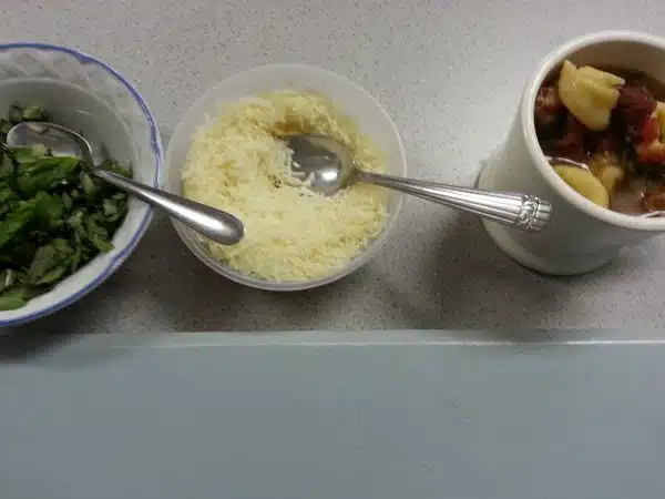 Three bowls are lined up on a counter containing chopped greens, grated cheese, and pasta in a tomato-based soup. Each bowl has a spoon.