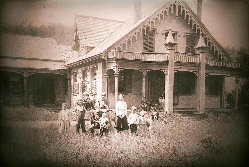 A vintage photo of a family and dog standing in front of a large Victorian-style house.