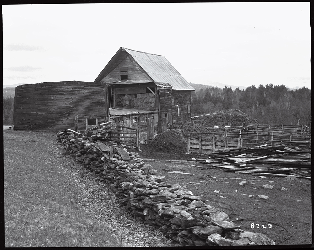 A weathered barn and silo stand beside a stone wall and wooden fencing on a rural farm landscape.