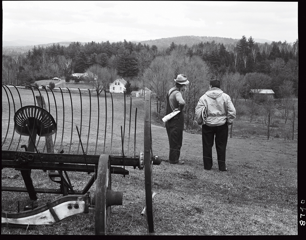 Two men stand in a rural field near old farm equipment, overlooking houses and trees in the distance.