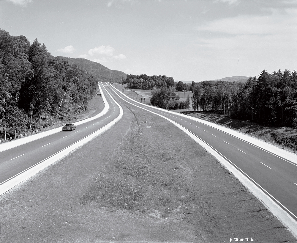A black-and-white photo of a mostly empty divided highway surrounded by trees and hills.