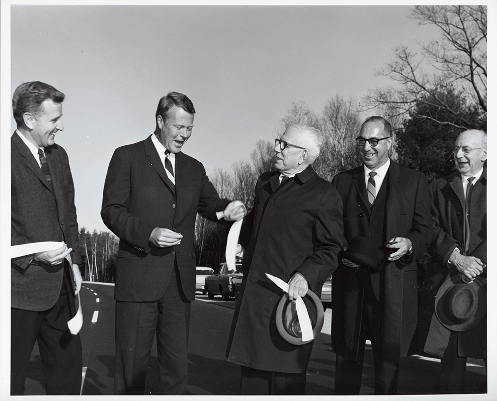 Five men in suits smiling and holding ribbon on a newly paved road, trees and cars in the background.