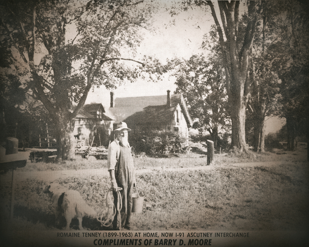 Romaine Tenney stands with a dog in front of a farmhouse and trees, holding a bucket and a walking stick.