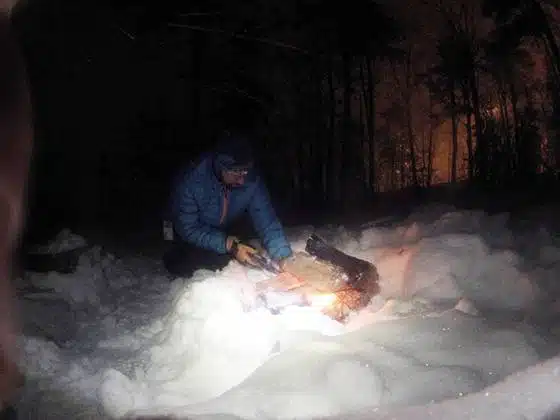 Person in a blue jacket tending to a small fire in the snow at night with trees in the background.