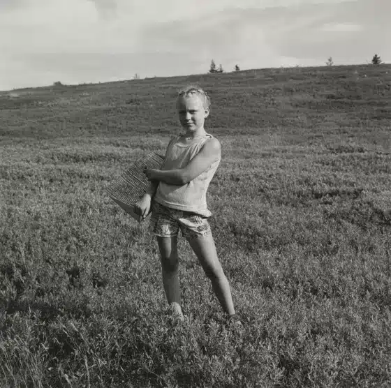 A young child stands in a grassy field holding a woven basket, with hills and a cloudy sky in the background.