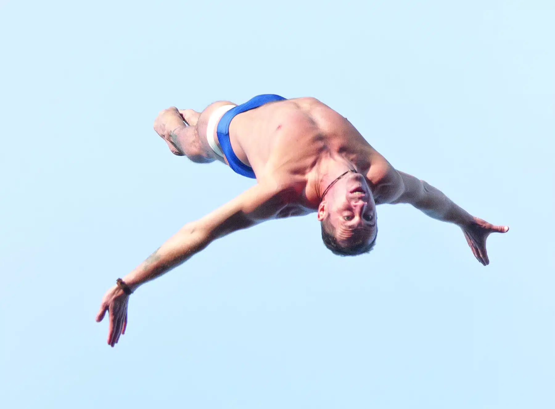 A man in blue swim trunks is mid-air, arms spread wide, executing a dive against a clear blue sky.