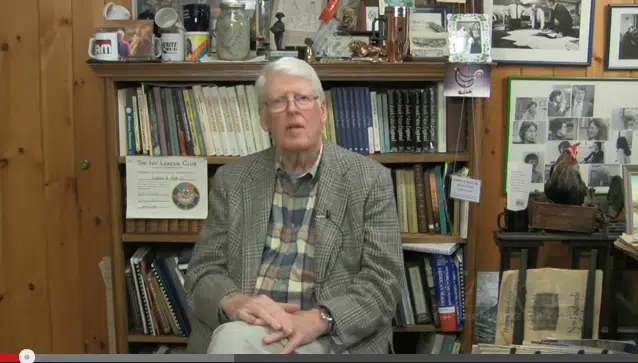 An elderly man with white hair and glasses sits in front of a bookshelf filled with books and various items. He is wearing a plaid shirt and a checkered blazer. Photographs and memorabilia decorate the background.