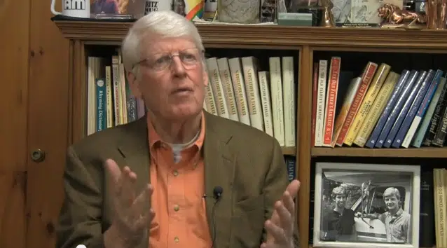 An elderly man with white hair and glasses, dressed in a brown blazer and orange shirt, gestures with his hands while speaking. He is seated in front of a bookshelf filled with books and a framed photo.