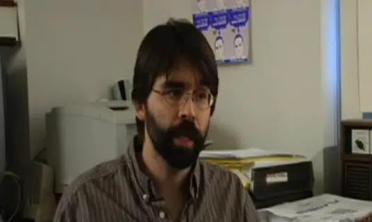 A man with glasses and a beard wearing a striped shirt is seated in an office with a printer, papers, and a bulletin board in the background.
