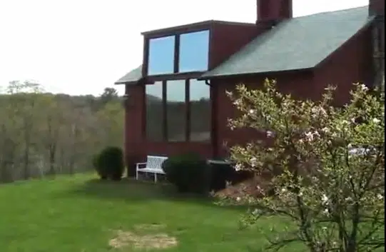 A brick house with large windows and a green lawn with some shrubs and a small tree in the foreground. There is also a white bench near the house.