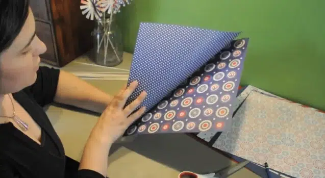 A person turning the pages of a book with patterned designs on each page, sitting at a desk with scissors and patterned paper nearby.