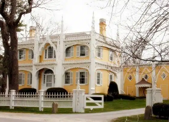 A two-story yellow house with white trim, ornate woodwork, and a white picket fence in front. The yard is kept neat with trimmed bushes and leafless trees.