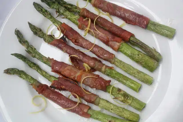 A plate of asparagus spears wrapped in crispy bacon, garnished with lemon zest.