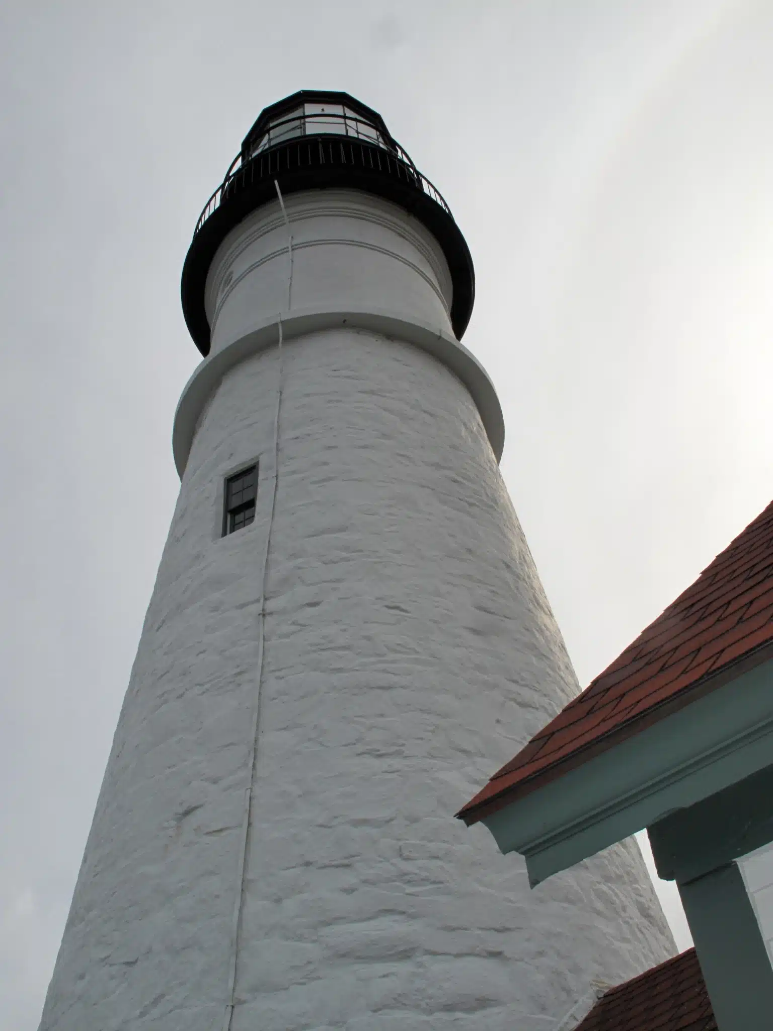 A tall white lighthouse with a black top stands next to a building with a red roof, viewed from below against a cloudy sky.