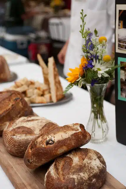 A display table with rustic loaves of bread, a plate of bread slices, and a vase with wildflowers. A person in a white shirt stands in the background.