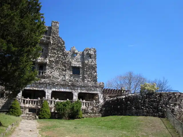 A stone building with three arches, surrounded by greenery and a clear blue sky. Stone steps and a partially bare tree are visible.