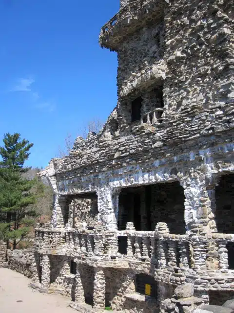 An old stone building with multiple levels and a weathered facade stands against a clear blue sky. Trees are visible in the background.