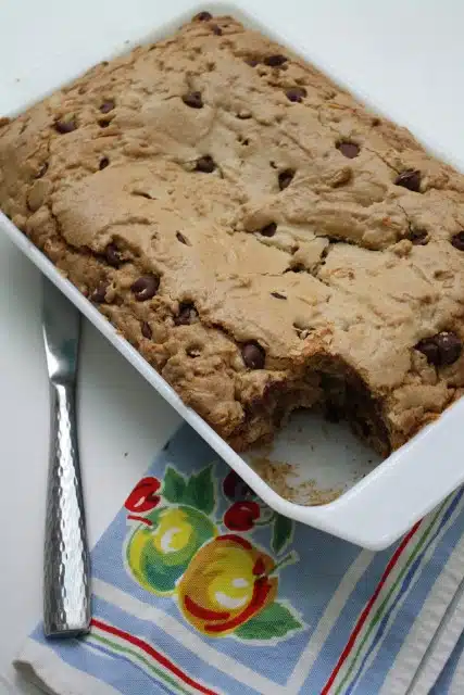 A baked chocolate chip cookie bar in a white rectangular dish with a piece missing, placed next to a silver knife and a blue cloth with fruit print and stripes.