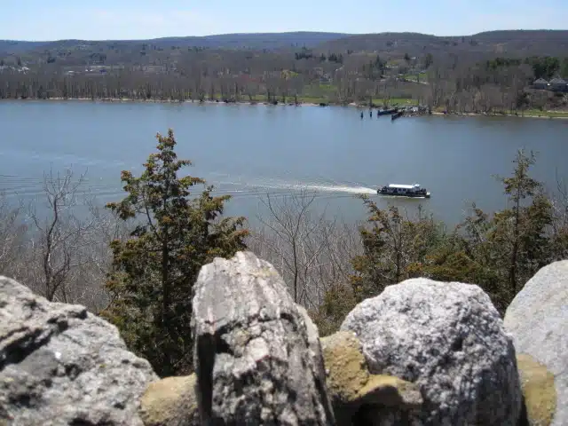 A small boat travels across a wide river with wooded hills and a few houses visible in the background. The foreground includes rocky terrain and leafless trees.
