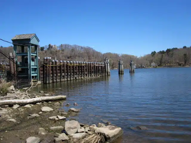 A river with a wooden pier structure extending into the water, a small blue building on the left, and trees and hills in the background under a clear blue sky.