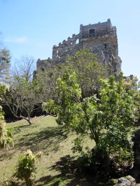 A stone castle ruin stands amidst greenery and trees on a sunny day with a clear blue sky in the background.