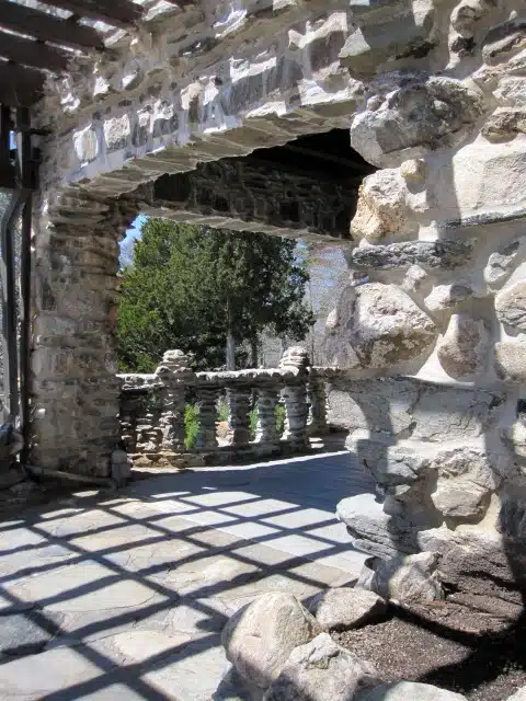 A stone archway leads to an outdoor balcony with a balustrade, casting shadows on the stone floor. Trees and clear sky are visible in the background.