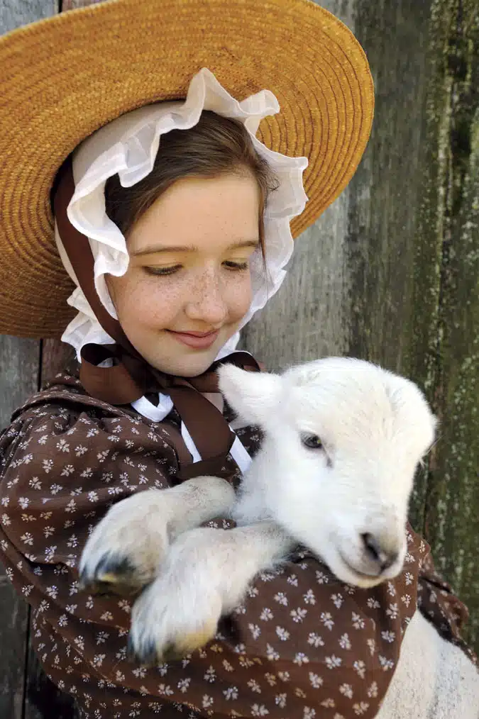 A girl wearing a brown bonnet and dress holds a white lamb close to her.