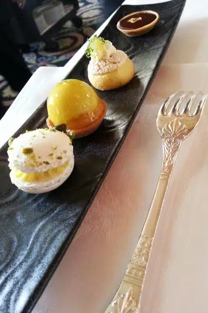 A rectangular black plate features four assorted elegant pastries and a decorative silver fork, placed on a white tablecloth.