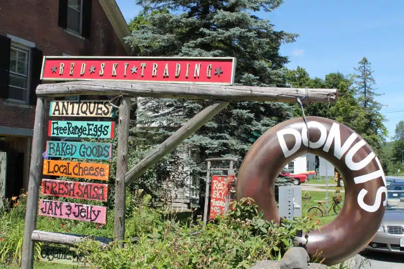 A rustic outdoor signboard for "Red Sky Trading" advertises antiques, free range eggs, baked goods, local cheese, fresh salsa, jam & jelly. A large donut replica hangs nearby. Trees and a car in the background.