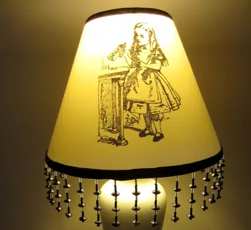 A lampshade with an illustration of a girl standing at a table. The edge of the lampshade is adorned with hanging beaded tassels.
