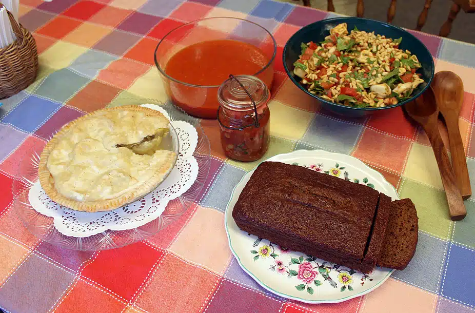 A table with a fruit pie, a jar of salsa, a bowl of tomato soup, a vegetable salad, and a loaf of bread on a floral plate.