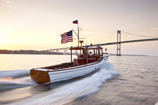 A motorboat with an American flag cruises through the water at sunset, with a large suspension bridge visible in the background.