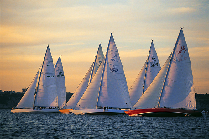 A fleet of 12-metre class yachts ("Intrepid", "Heritage", "American Eagle", "Weatherly", "Nefertiti", and "Columbia"), all former Cup contenders, race in tight formation in Narragansett Bay.